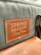 A brown leather patch with stitched edges and embossed text reading ’spring meadow dairy farm’ is affixed to a dark gray canvas bag.