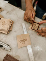 Light brown leather square with a laser-engraved design of a stylized bird or mythical creature in dark brown, framed by an engraved border.