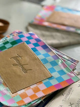 A vibrant, iridescent checkered wallet with a tan leather patch featuring the brand name ’thandana’ embossed in cursive script.