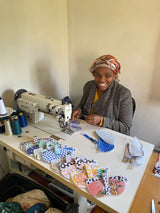 A woman in a patterned headscarf and tweed jacket sits at a white zote industrial sewing machine, surrounded by colorful fabric pieces on the table.