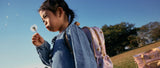 A young girl in a denim dress blows on a dandelion puffball.